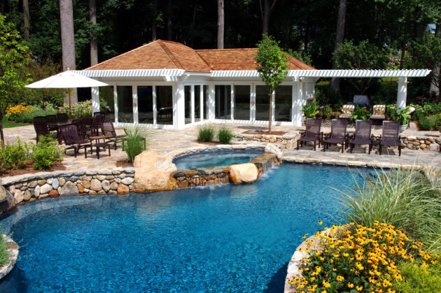 A luxury backyard in Massachusetts featuring a custom pool, spa, stone patio, elegant pool house, and a white pergola for shade.